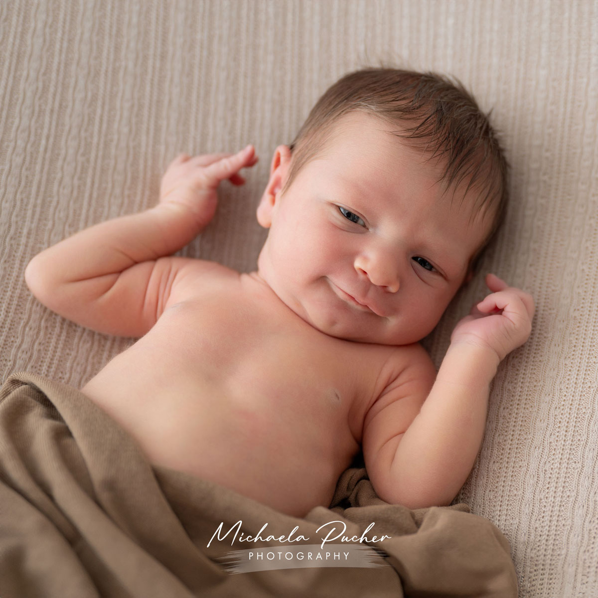 Neugeborenes liegt auf einer weichen, beige strukturierten Decke und schaut wach mit einem feinen, charmanten Lächeln in die Kamera.